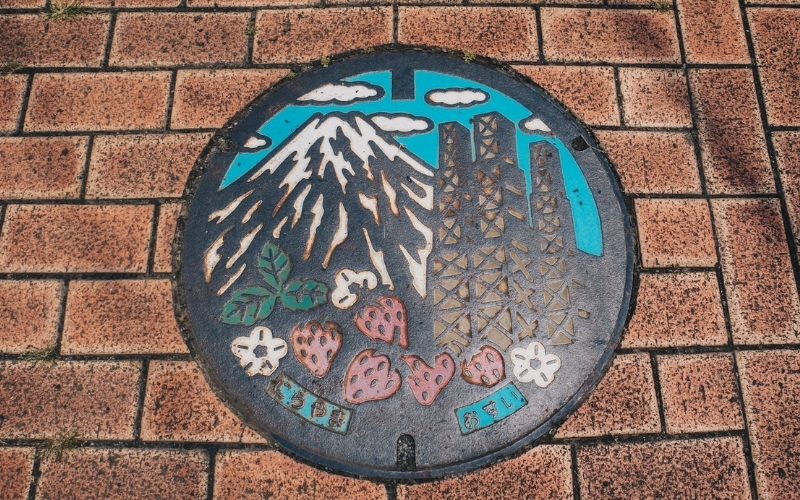 Japanese manhole which features an illustration of a mountain, a building, and strawberries